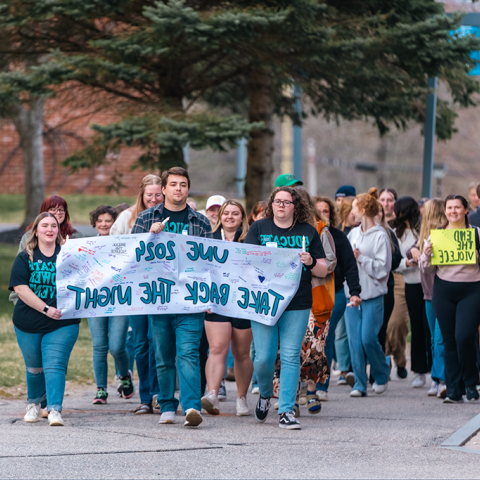 A group of students marches on campus holding a sign that reads "UNE 2024 Take Back the Night"